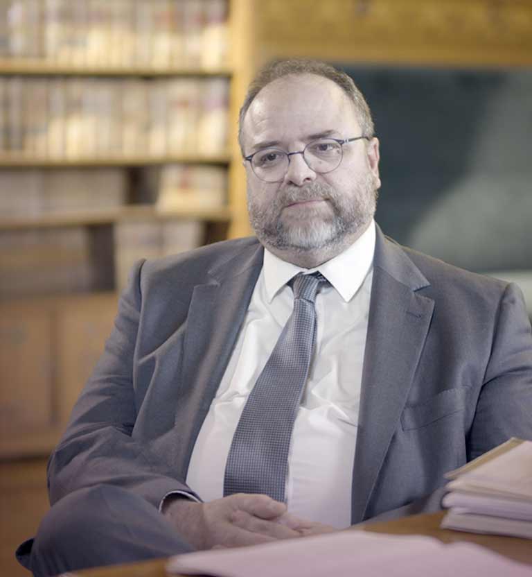 Hombre adulto sentado con gafas, anteojos, vistiendo un traje con corbata en una biblioteca