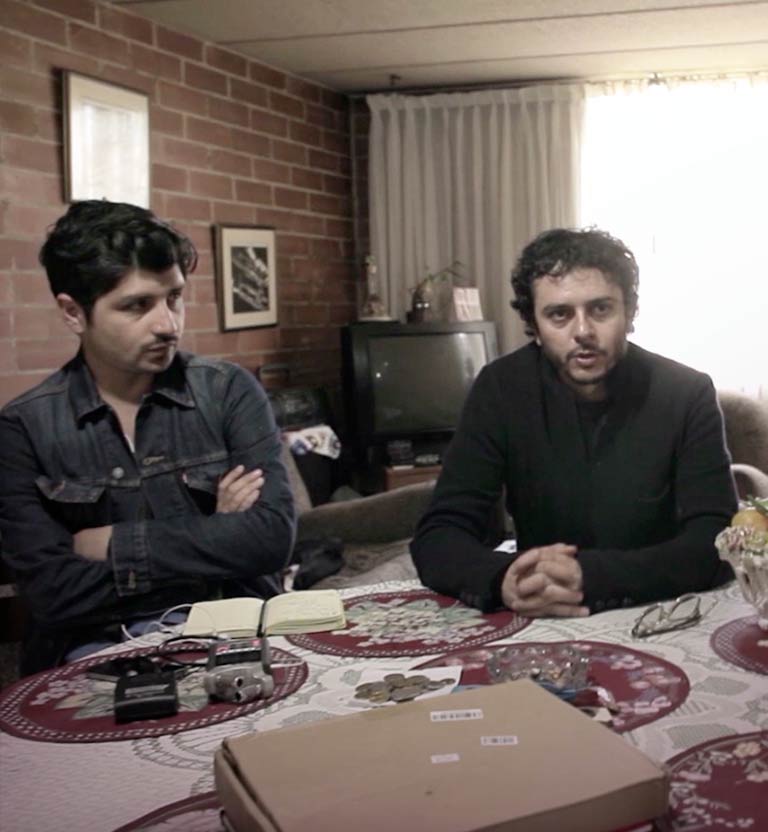 Dos jóvenes sentados en un comedor dentro de un apartamento. Al fondo una sala.