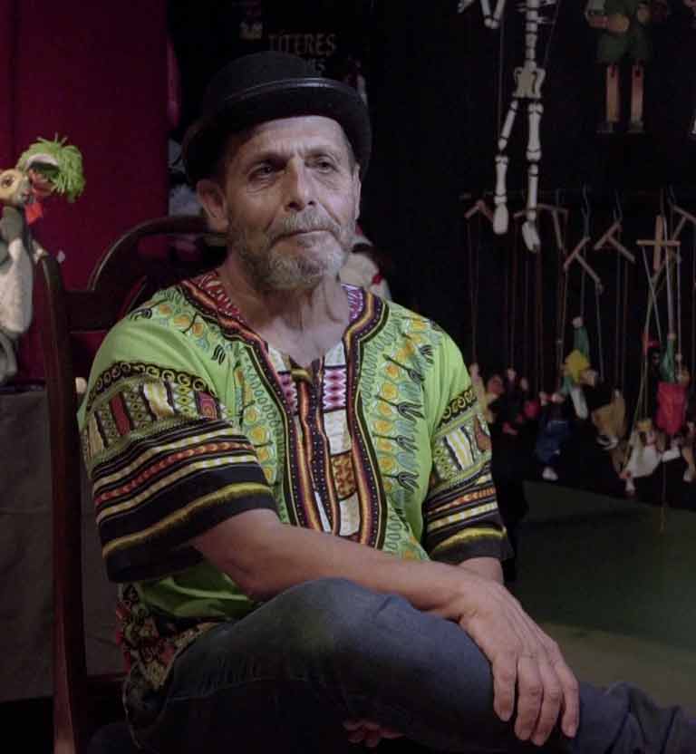adulto mayor sentado con unas marionetas detrás suyo