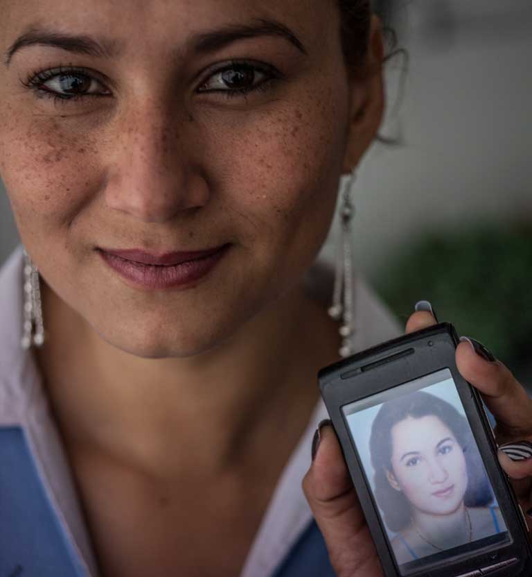 Mujer joven sonriendo, sostiene en su mano un celular con una fotografía de ella misma más joven