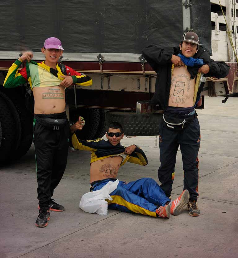 Tres hinchas de fútbol jóvenes con la camiseta arriba mostrando su tatuaje al lado de un camión sobre la calle.