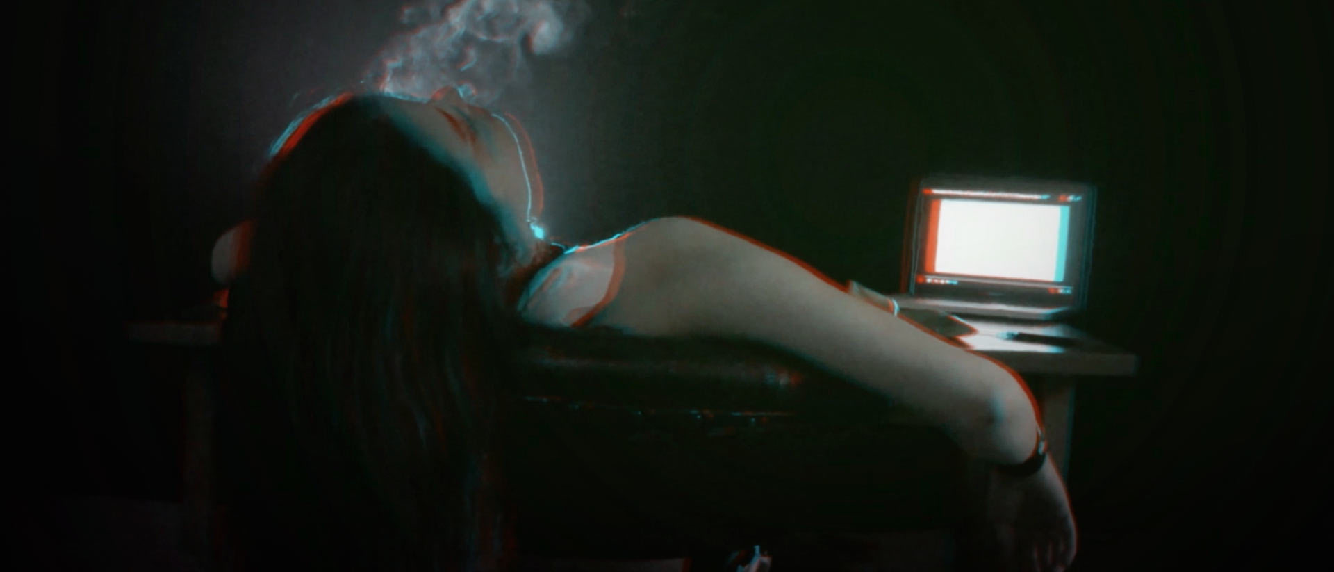 Mujer desgonzada en una silla con la cabeza colgando, con una pantalla de computador al fondo en un cuarto oscuro.