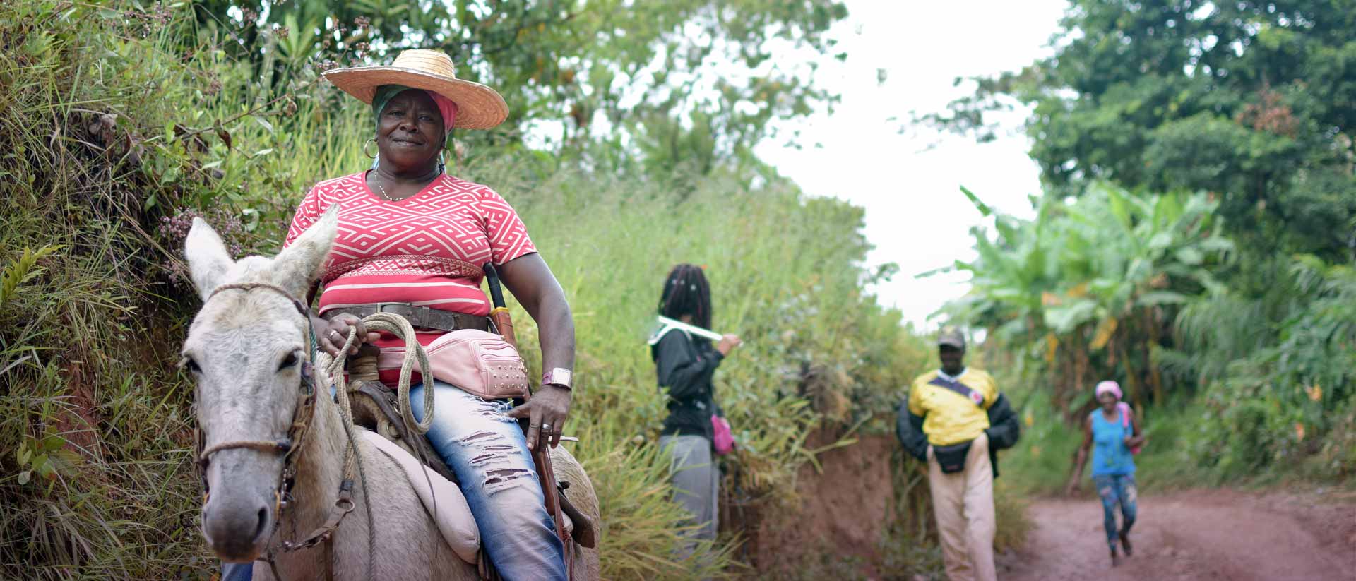 mujer afro adulta sonriendo, montando a caballo junto a personas afro caminando por una vereda