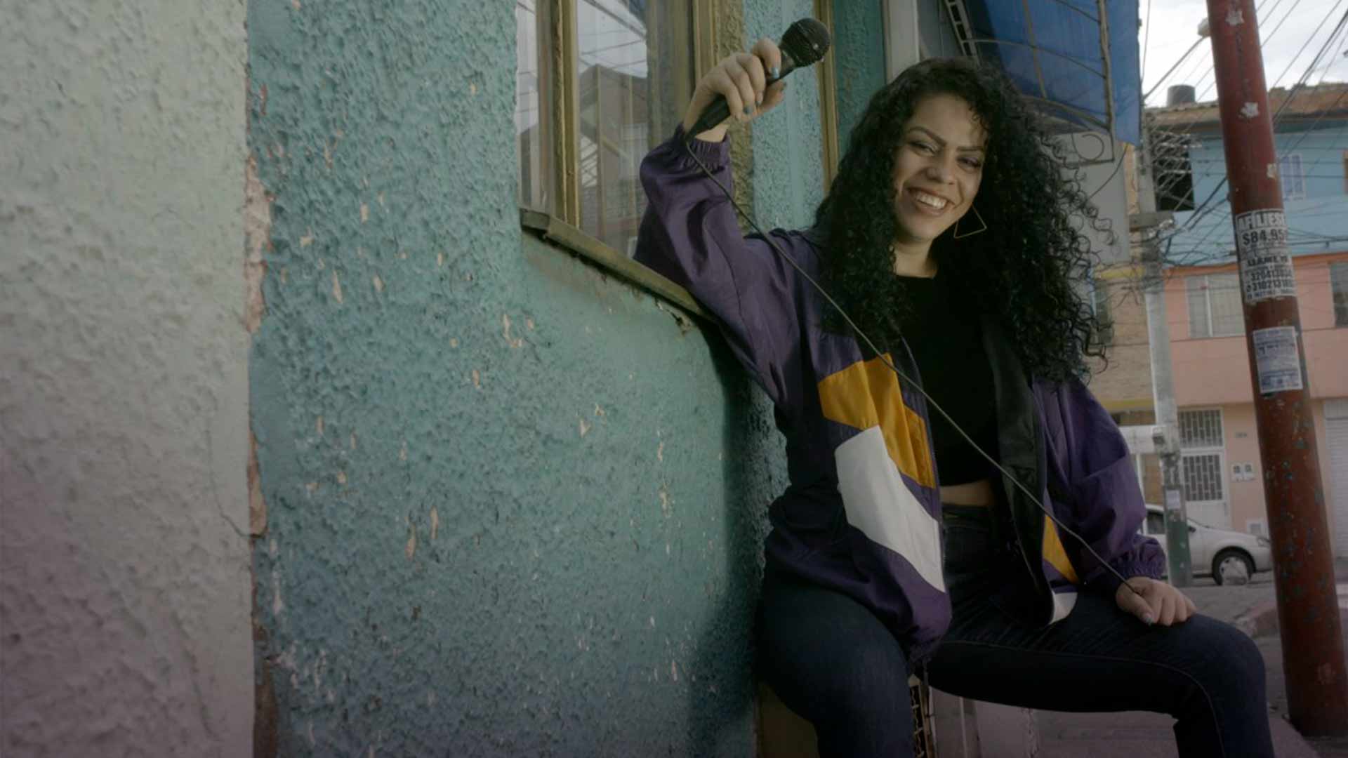 Mujer joven apoyada en una pared con un micrófono en la mano.