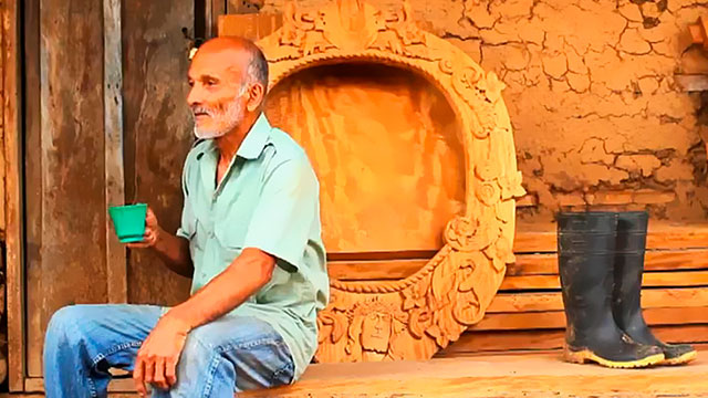 Campesino sentado al lado de una pieza tallada en madera. Al fondo una casa en barro. 