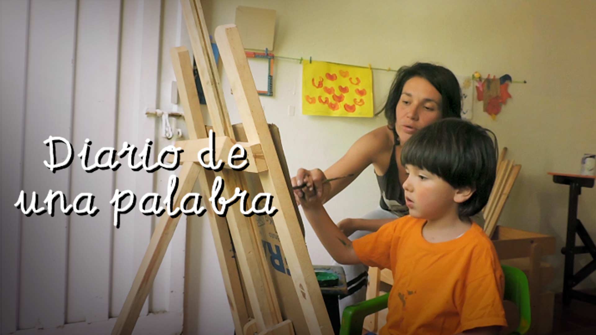 niño de pie pintando un cuadro junto a su madre con algunas pinturas detrás